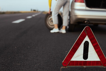 a girl in casual clothes stands near the rear wheel of the car and rolls the spare wheel to change. Car breakdown, the wheel has been pierced. Close-up and focus on the emergency stop sign,