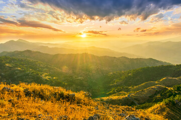 Obraz premium Mountain valley during sunset or sunrise. Rocks and slopes covered with green trees with amazing cloudy sunny sky on background.