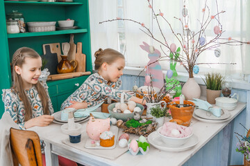 Girls, sisters at table with Easter decoration. Celebration in kitchen.Tablescape for home Easter holiday.Family religious traditional festive food, meal. Colored eggs,cake, funny bunny, candy sweet