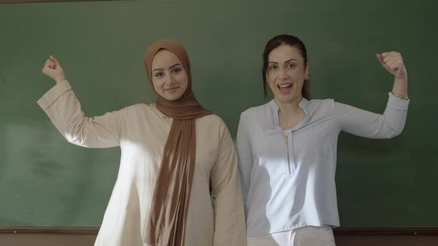 Female Teacher In Hijab And Female Colleague Looking At Camera In Classroom With Arms In Front Of Blackboard. Successful Teachers Smile And Raise Their Arms To Show Their Biceps.