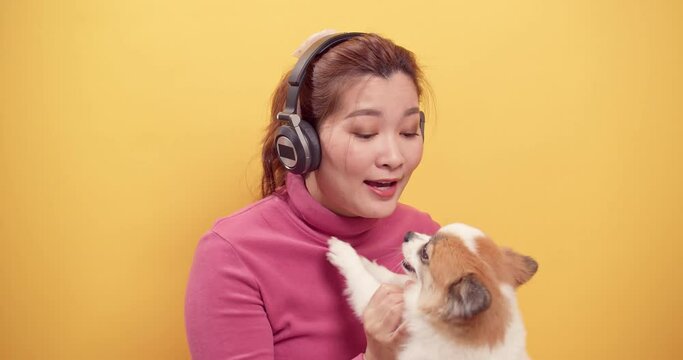 Asian gorgeous woman uses digital tablet and streaming application for happy listening to music on headphones while with playing the dog for relaxation on bright yellow background. Relaxation concept.
