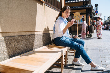 Obraz na płótnie Canvas asian girl in casual wear is checking phone messages while taking a rest sit on the wooden bench at hanamikoji street in gion district, Kyoto japan with sunlight