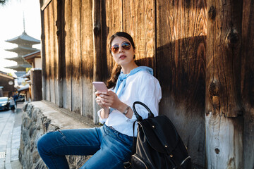 curious trendy Korean female holding phone is looking into distance while resting by a traditional wooden building near Yasaka pagoda in Kyoto, japan.
