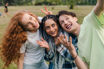 Three teenagers taking selfie for posting in internet, standing in the summer park. Vloggers taking a video. Trendy happy mixed people.