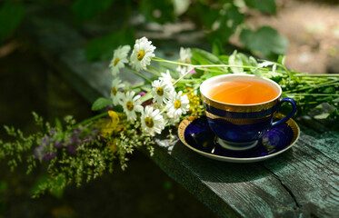 Cup of chamomile herbal tea and chamomile flowers and wildflowers on wooden table in garden, blurred background, soft focus