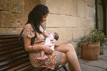 young mom breastfeeding newborn baby daughter in public outdoors in the street on a bench