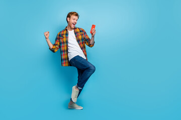 Full size photo of stylish cheerful funky guy with red hair dressed plaid shirt hold smartphone fist isolated on blue color background