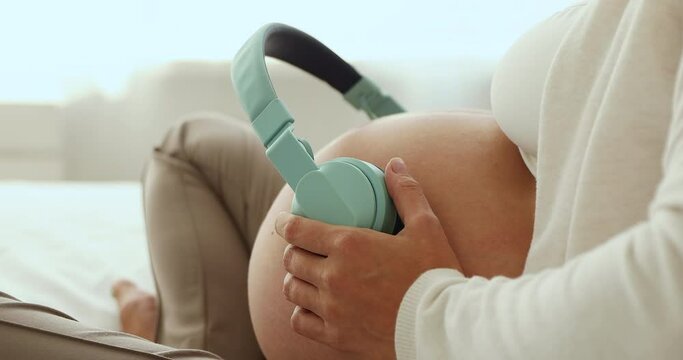 Close up, unknown pregnant woman put headphones on belly, play classical tune for prenatal child development, enjoy carefree rest at home. Pregnancy music for expectant mother and unborn baby concept