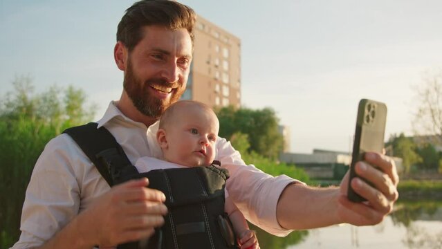 Smiling happy young father with a little newborn baby in a slings carrier. Use phone to take selfies on street near lake in park at sunlight. Ergo backpack. Parents. Gadget. Smartphone. Cell. Slow