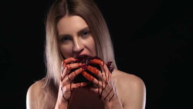 Young Attractive Blonde Woman Eating A Heart, Blood Dripping From Her Hands. Hungry Girl Ripping Raw Red Meat With Her Teeth, Chewing, Looking At Camera. Studio Portrait Shot. Vampire, Horror Concept