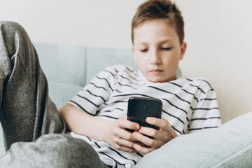 Teenage boy sitting on couch using smartphone and playing video games on internet online at cozy home. Child holding mobile phone and looking at screen. Social media