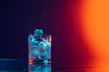Ice cube falls into a transparent glass of water standing on mirror surface over gradient black...