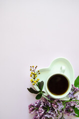 Flatlay with a white cup of coffee and a saucer in the shape of a heart in the lower corner of a light purple background, large copyspace and lilac flowers, small yellow flowers and dark green leaves