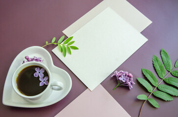 close-up diagonal Mockup of white cards and a pink envelope on a dark purple background with leaves and lilac flowers with a coffee cup and heart-shaped saucer