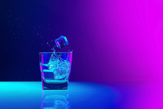 Ice cube falls into a transparent glass of water standing on mirror surface over gradient blue-pink background in neon light. Art, beauty, drinks