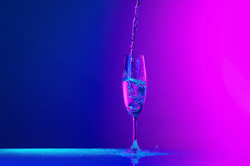 Authentic hand pouring pure water splash from wine or champagne glass over bright blue-pink gradient background in neon light. Art, drinks, creativity