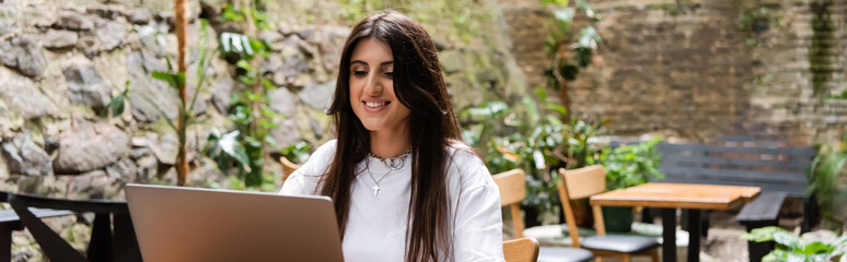 Cheerful brunette freelancer using laptop on terrace of cafe, banner