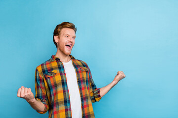 Photo of cheerful handsome satisfied man with red hair dressed checkered shirt hold fists up shout yes isolated on blue color background