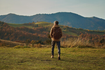 Back view tourist adventurous man with backpack stands on mountain meadow and enjoys inspiring epic landscape over mountains