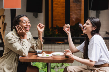Laughing multiethnic girlfriends talking near coffee and desserts on cafe terrace