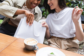 Cropped view of multiethnic girlfriends holding shopping bag near coffee in outdoor cafe