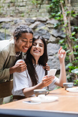 Positive african american woman hugging friend with smartphone near cups of coffee in outdoor cafe