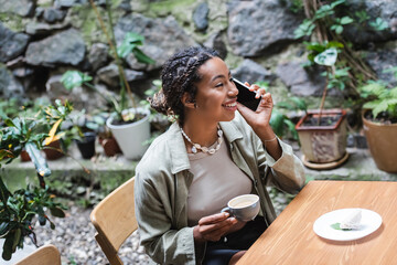 Cheerful african american woman talking on smartphone and holding cup of cappuccino in outdoor cafe