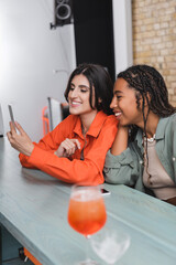 Multiethnic girlfriends using smartphone near cocktail in cafe