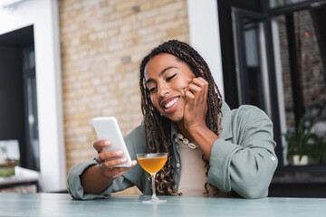 Positive african american woman using mobile phone near cocktail in cafe