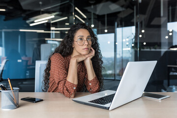Young beautiful hispanic woman with curly hair and glasses, working in modern office using laptop, female worker thinking and sad, business woman upset