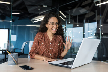 Plakat Successful and beautiful hispanic woman working inside modern office building, businesswoman using laptop for video call smiling and waving, greeting gesture, online conference with colleagues
