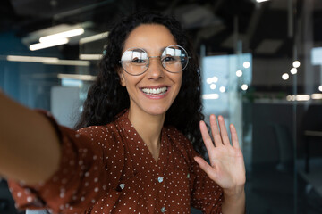 Beautiful business woman with curly hair wearing looking at smartphone camera, waving her hand in greeting gesture, talking with friends and colleagues, using video call app
