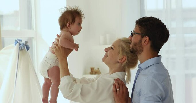 Parents spend priceless time with adorable newborn baby, couple standing in modern nursery play with daughter in diaper. Family bond, unconditional love, happy parenthood, carefree babyhood concept
