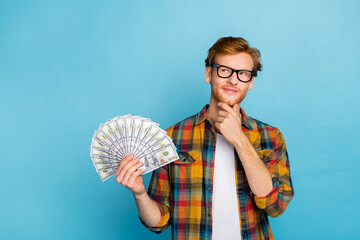 Photo of minded cheerful guy dressed checkered shirt look red hair empty space hand on chin hold money isolated on blue color background