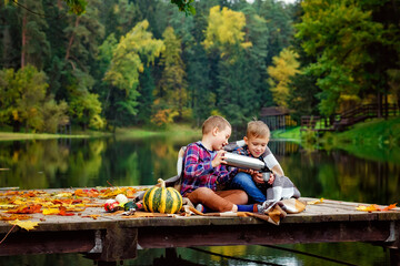 A boy pours tea from a thermos by the lake on the pier. Friends on a picnic in nature in autumn....
