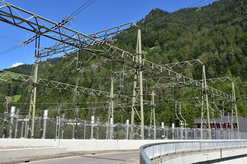 Fototapeta na wymiar Electric poles with overhead cables, expulsion fuses, surge arrester and other components as a part of hydroelectric power plant on reservoir Mapraggsee. In background is green hill with trees.