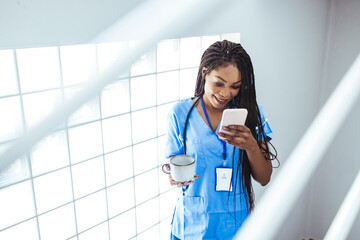 Portrait of young woman doctor in  scrubs sitting while using smartphone in hospital, relax after...