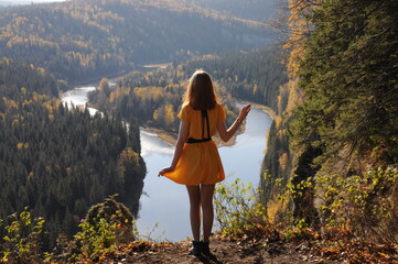 Rear view of young traveler in yellow dress, who stands on edge of rocky wall against backdrop of...