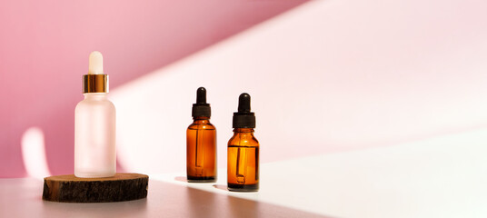 Banner of three transparent amber and frozen glass dropper bottles on pink background in the sunlight. Wooden podium. Pipette with fluid hyaluronic acid, serum, retinol. Cosmetics, healthcare, beauty 