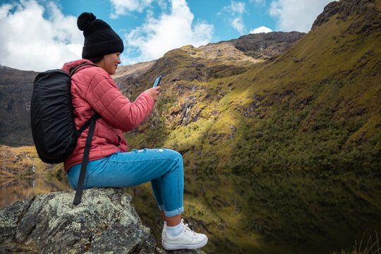 Excursionista atractivo sentado y mirando su teléfono junto a un lago tranquilo en las hermosas montañas