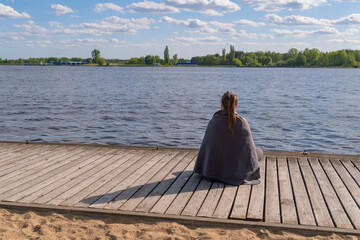 a woman sits on the edge of a pier made of wooden bars, a woman covered with a warm gray blanket looks into the distance at the blue water of the lake on the other shore, a forest strip, sunny, windy 