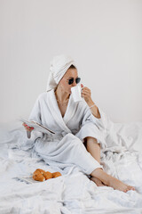 Happy woman in a white bathrobe and with a towel on her head drinks coffee and eats a croissant in...
