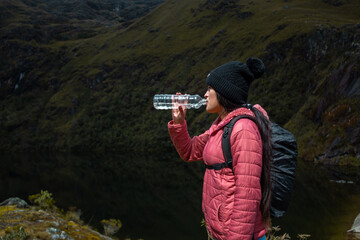Fototapeta na wymiar Caminante en una chaqueta rosa bebiendo agua junto a un lago tranquilo rodeado de montañas cubiertas de musgo