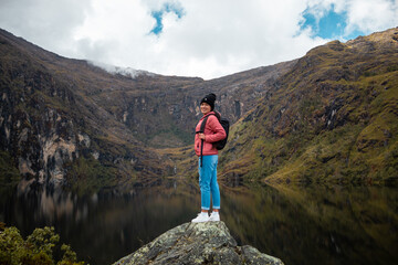 Una chica con una chaqueta rosa y mochila parada  junto a un tranquilo lago en las hermosas montañas - obrazy, fototapety, plakaty
