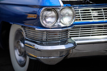 The front quarter panel, headlight housings, and chrome grill and bumper, and white wall tire of a vintage classic blue auto.