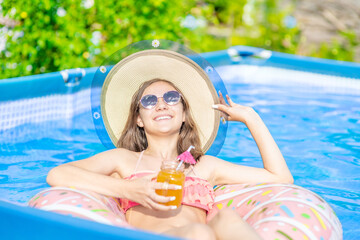 Happy teen girl wearing sunglasses and summer hat drinks orange cocktail on colorful inflatable ring in outdoor pool at hot summer day