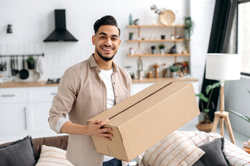 Joyful modern indian or arabian guy, stand at home in the living room, holding a large cardboard...