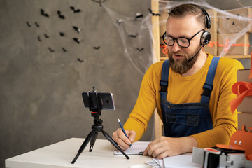 bearded man in glasses business owner sits in halloween decorated home office. Writes with a pen on paper. selling goods in honor of the holiday of all saints, discounts and promotions.