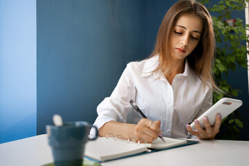 beautiful young businesswoman workingat the cafe indoors, using mobile phone while making notes