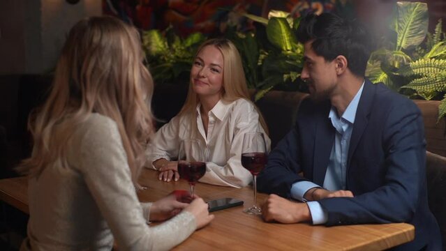 Hand held shot of two cheerful blonde women and handsome man in suit sitting in restaurant, drinking wine and talking laughing having fun. Business partners having drink at hotel bar, slow motion.